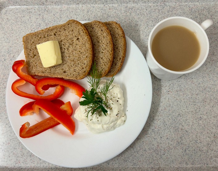 Na zdjęciu znajduje się Kawa z mlekiem,  Chleb graham, Masło extra 82%, Serek homo o smaku naturalnym z ziołami, papryka świeża